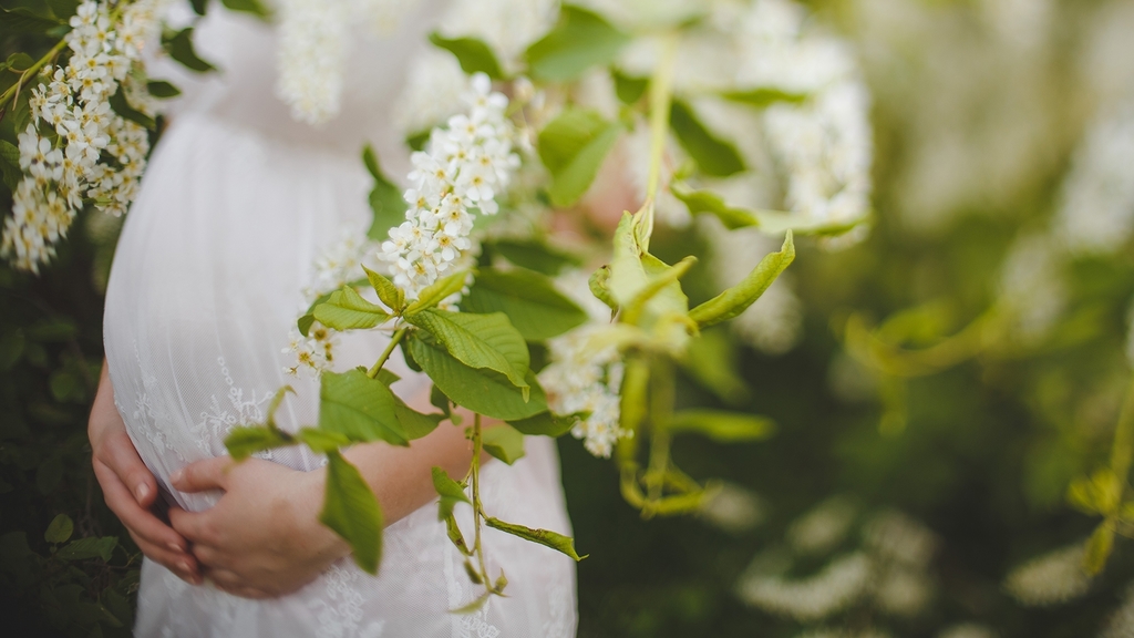 Vergrößerung des Bildes für Schwangere Frau im weißen Kleid; sie steht hinter einer grünen Pflanze mit weißen Blüten.
