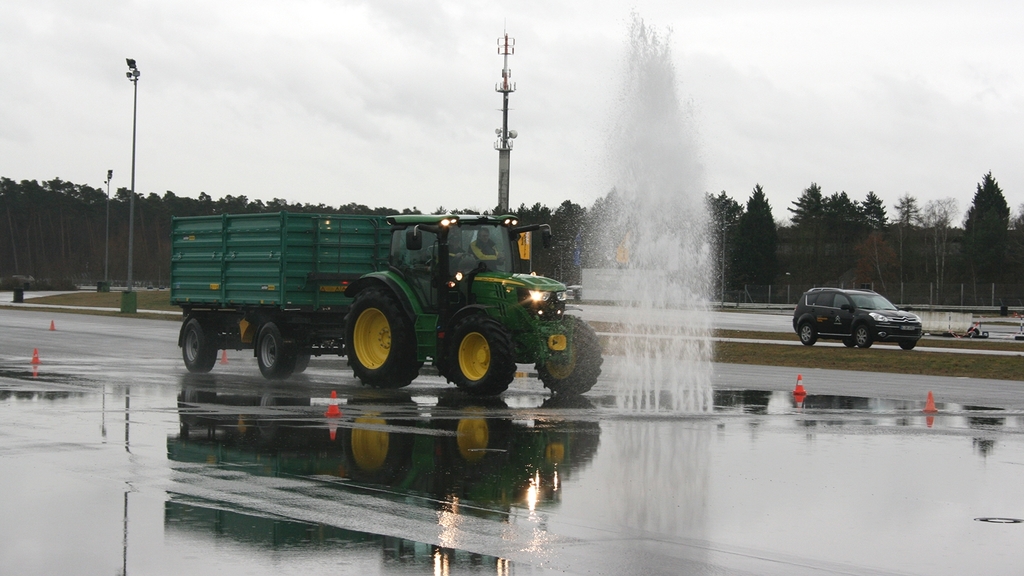 Vergrößerung des Bildes für Traktor mit Anhänger fährt auf einer nassen Fahrbahn durch die Wasserfontäne.