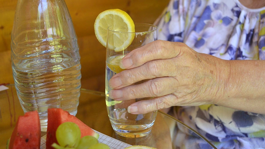 Vergrößerung des Bildes für Detailfoto zeigt den Arm einer älteren Frau mit einem Glas Wasser in der Hand. Das Glas Wasser ist mit einer Zitronenscheibe dekoriert..