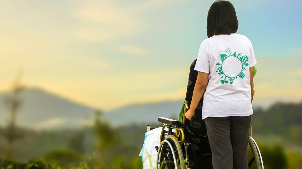 Vergrößerung des Bildes für Rückenansicht einer jungen Frau, die - mit einem Patienten im Rollstuhl in der Natur stehend - die Aussicht auf eine Berglandschaft genießt.